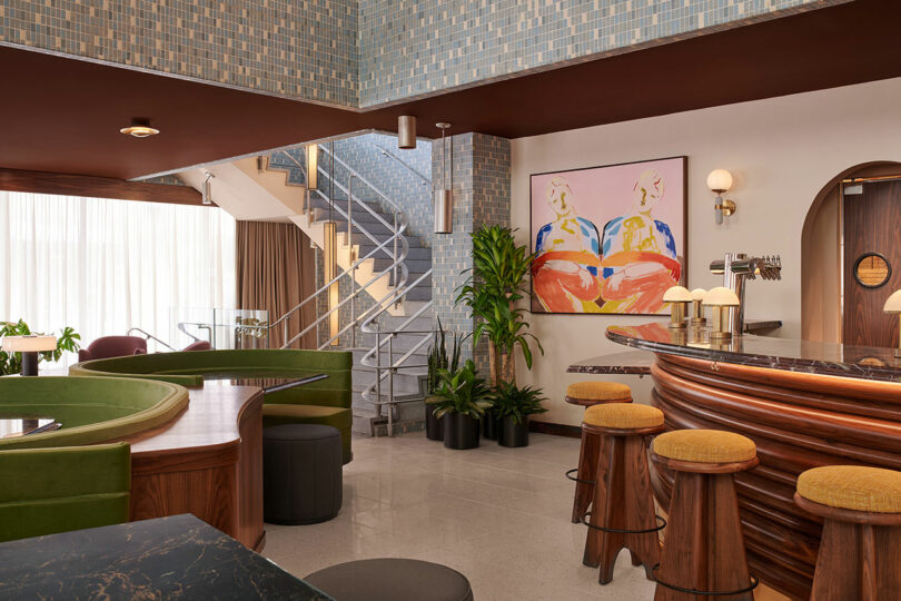 Modern lounge with curved green seating, wood-accented bar and stools, indoor plants, a spiral staircase, and colorful abstract artwork on the wall. Natural light flows through large windows in this Municipal Grand-inspired setting.