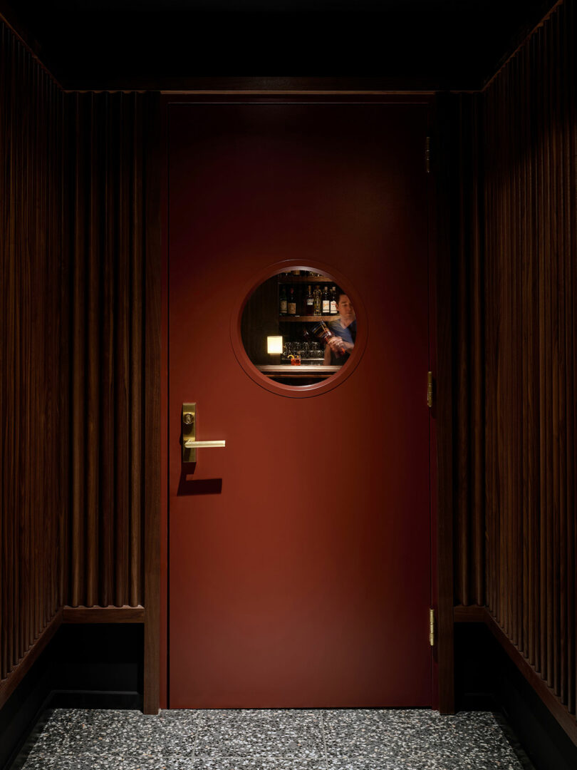 A round window in a closed red door reveals a person behind the bar at Municipal Grand, surrounded by shelves of bottles in a dimly lit interior with wood-paneled walls.