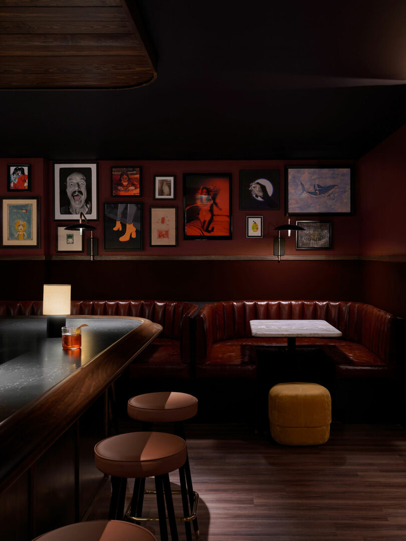 A dimly lit bar at the Municipal Grand with a marble counter, round stools, red leather booths, a yellow ottoman, and framed artwork on dark red walls. A drink and a lamp sit on the bar.