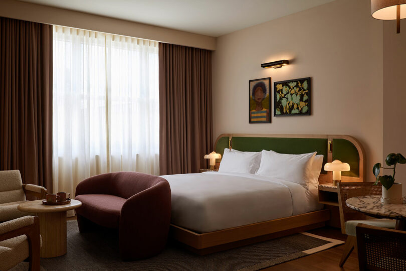 A modern Municipal Grand hotel room with a double bed, a curved burgundy sofa, two armchairs, a small round table, and framed artworks above the bed. Natural light enters through a large window.
