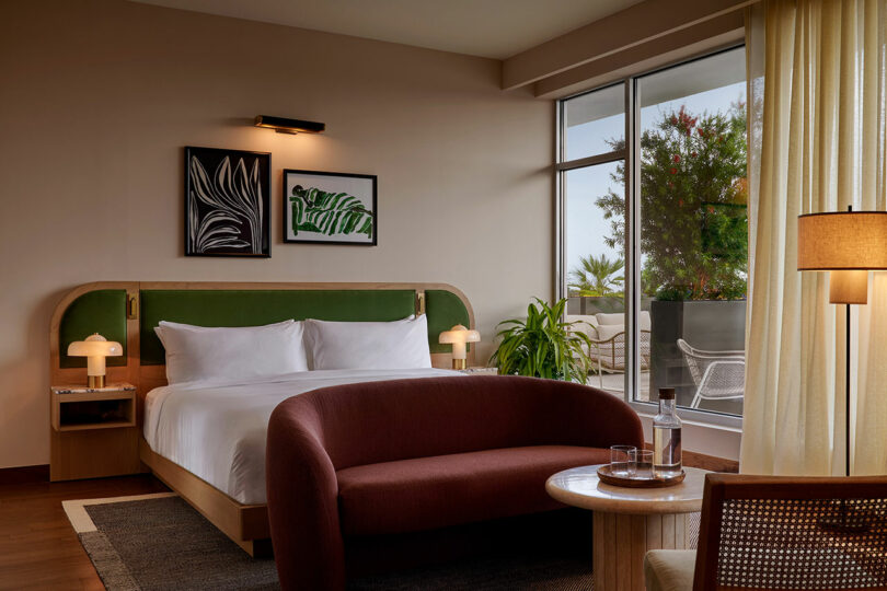 Modern Municipal Grand hotel room with a double bed, green headboard, two framed leaf prints, maroon curved sofa, round table with a water bottle, large window, and balcony view.