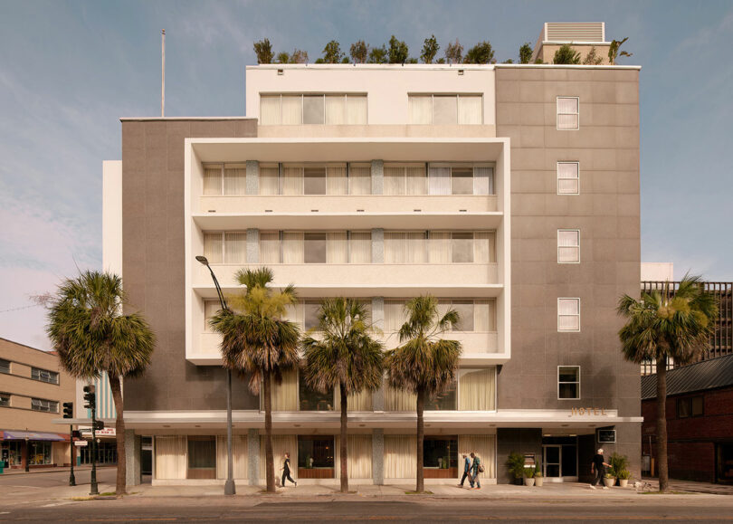 A modern mid-rise hotel building, the Municipal Grand boasts a gray and white facade, balconies, rooftop greenery, and palm trees in front; several people stroll along the sidewalk.