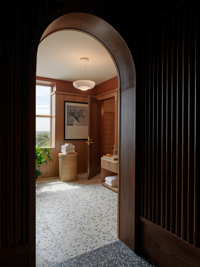 View through an arched doorway into a bathroom with terrazzo flooring, wooden walls, a potted plant, stacked towels, and framed artwork—bathed in natural light with a touch of Municipal Grand charm.