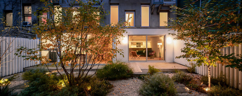 Modern townhouse backyard at dusk, with illuminated windows, sliding glass doors, garden landscaping, trees, and gravel paths visible.