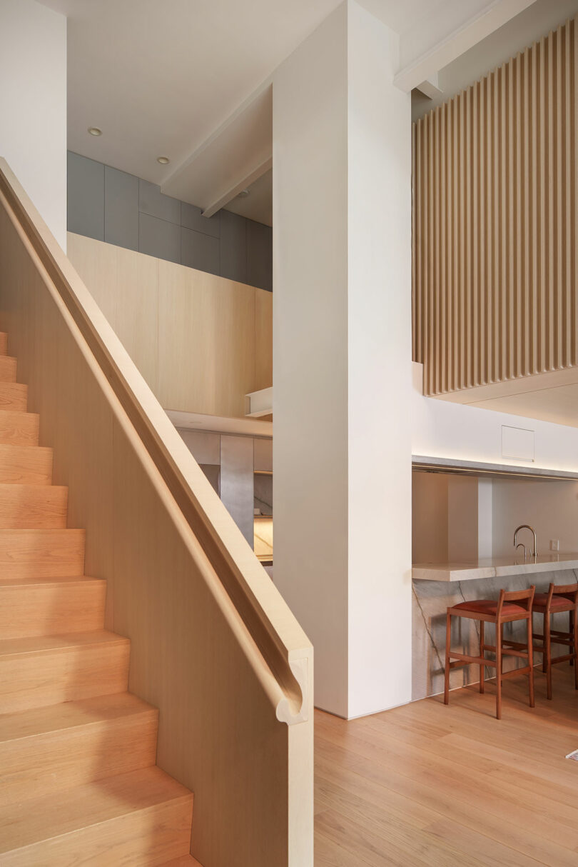 Modern interior with light wood staircase, open kitchen area with bar stools, and vertical wooden slats as room divider. Neutral tones and minimalist design elements throughout.