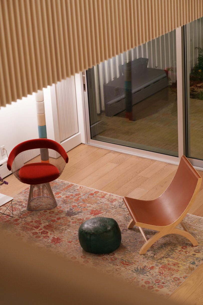 Modern living room with a red chair, tan leather lounge chair, green pouf, and patterned rug near sliding glass doors and hardwood floors, viewed from above.