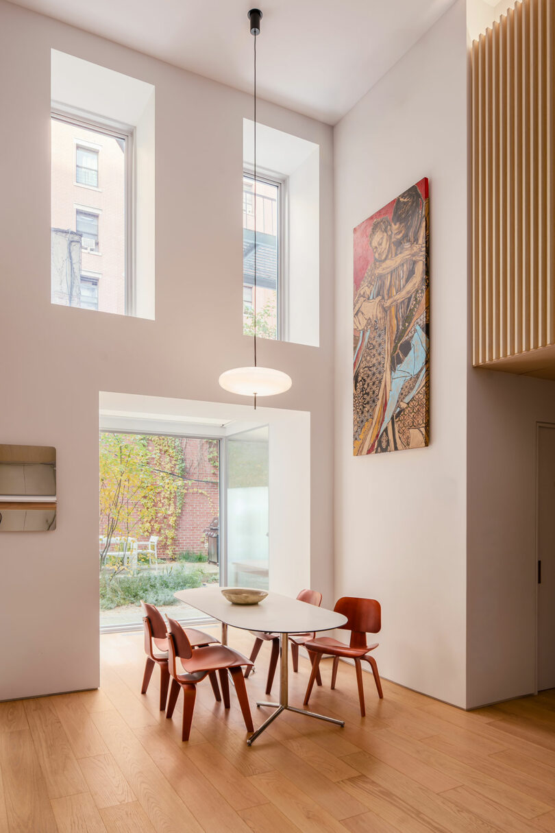 A modern dining area with a white table, four red chairs, large windows, a pendant light, wood flooring, and a colorful painting on the wall. A garden is visible outside.