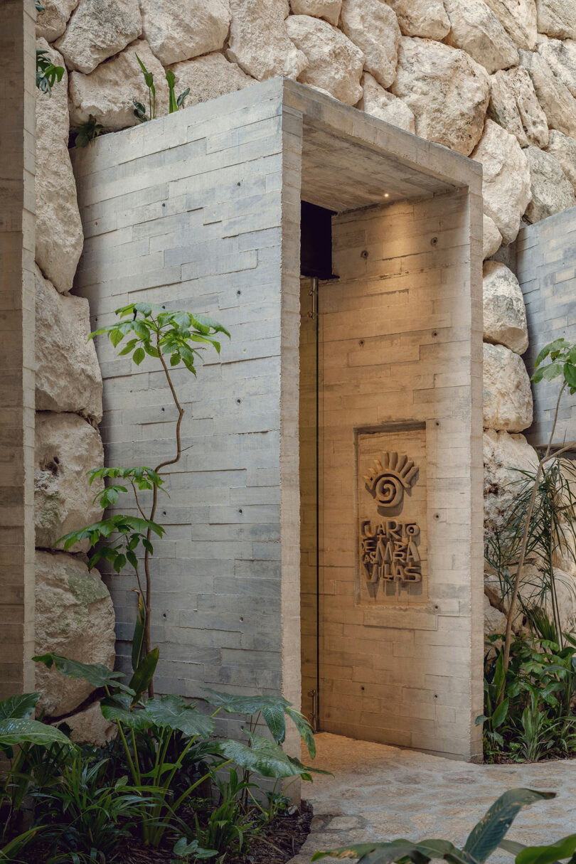 A modern stone entryway with a sign reading "Carden Mar Villas," surrounded by lush plants and a wall of large, rough stones.
