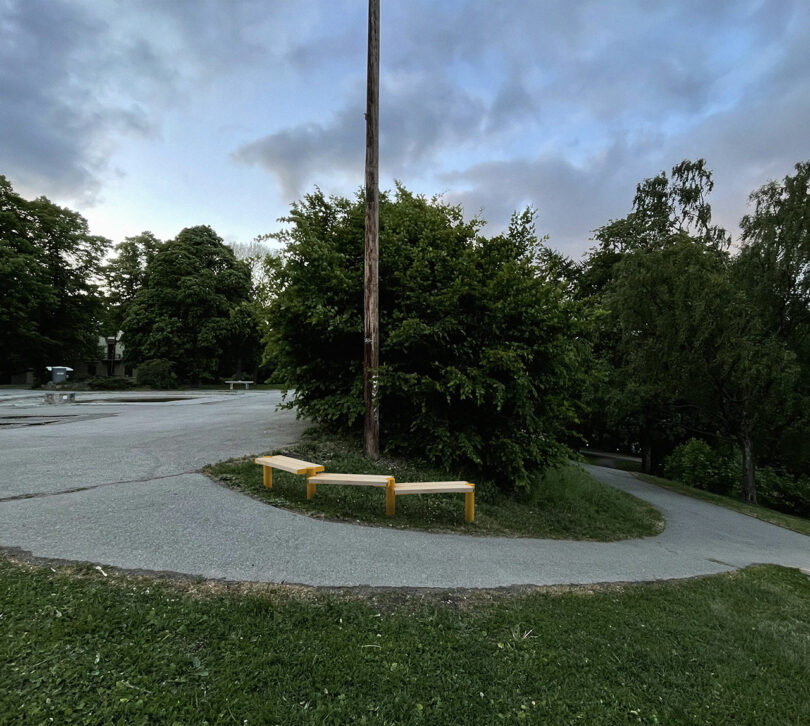 A paved path curves around a bush with a wooden pole, two benches beside it, and trees in the background under a cloudy sky.