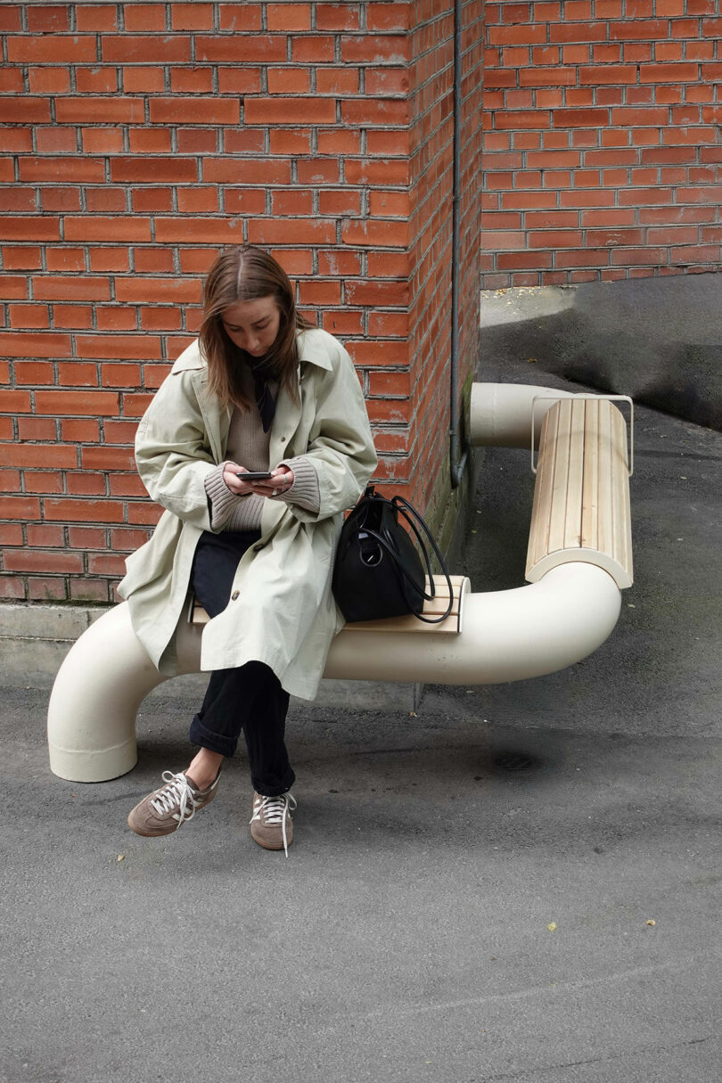 A woman in a light trench coat sits on a curved bench by a brick wall, looking at her phone with a black bag beside her.