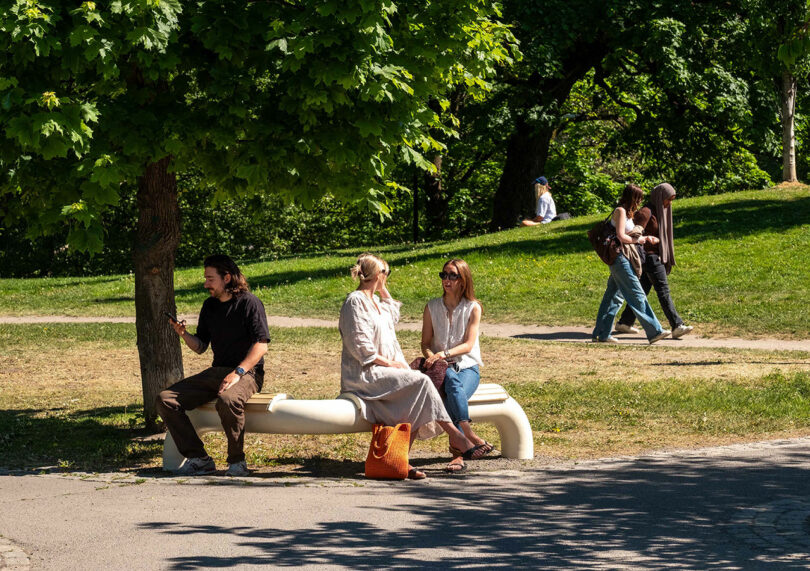 Three people sit on a curved bench under a tree in a park, while two pairs of people walk along paths in the background on a sunny day.