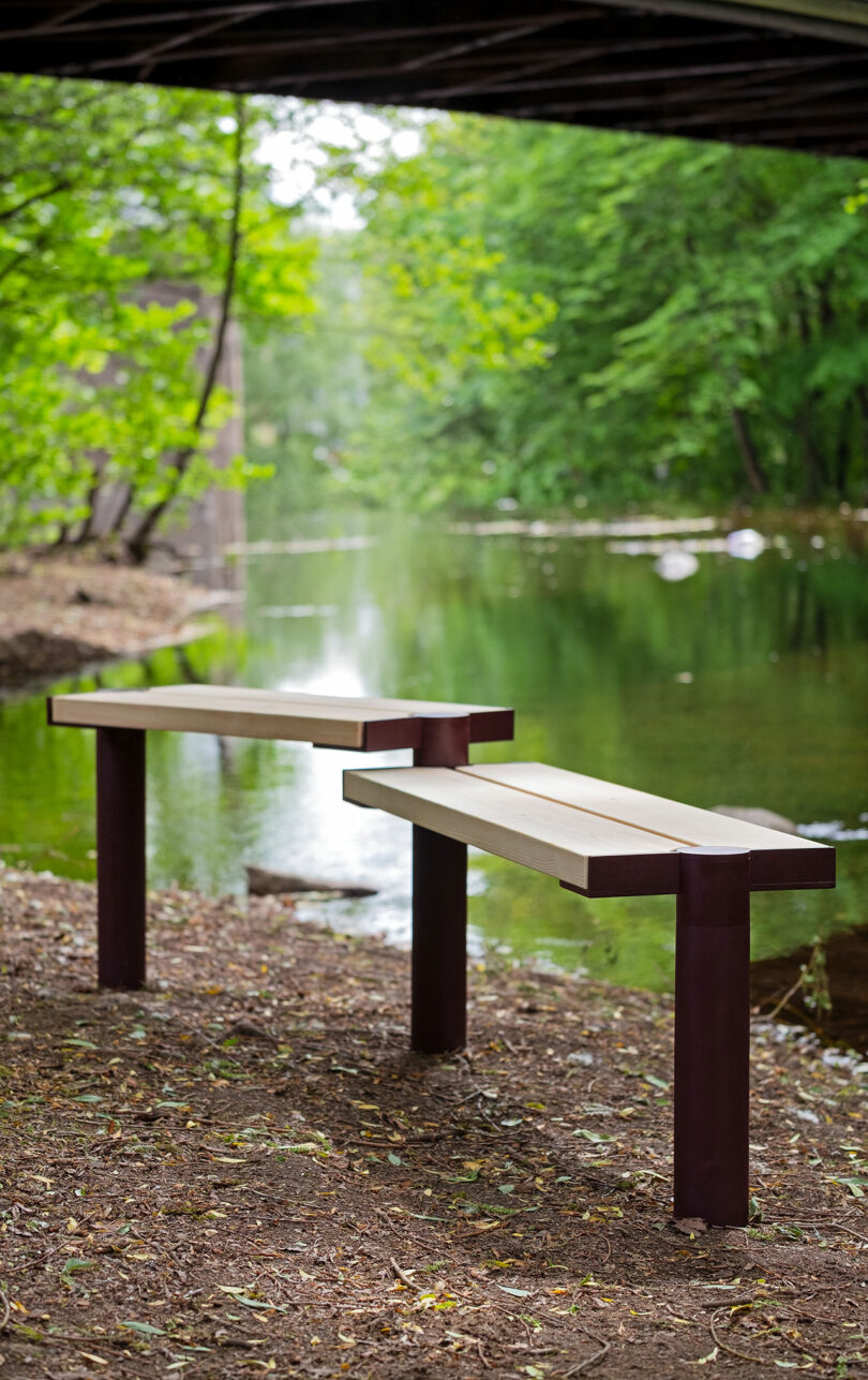 Two modern benches with wooden seats and metal legs are placed on the ground near a calm river, with green trees and a bridge overhead in the background.