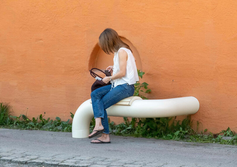 A woman sits on a curved, pipe-shaped bench against an orange wall, looking into her bag.