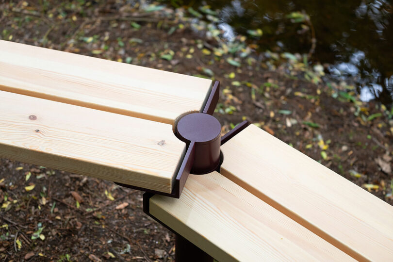 A close-up of two wooden benches joined at a right angle by a metal connector, positioned outdoors near soil and a body of water.