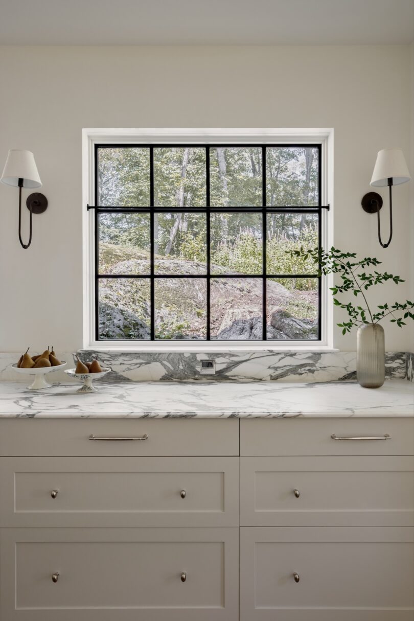 A kitchen counter with a marble surface, a vase with green branches, a bowl of pears, white drawers, and a window with black grid framing an outdoor view.