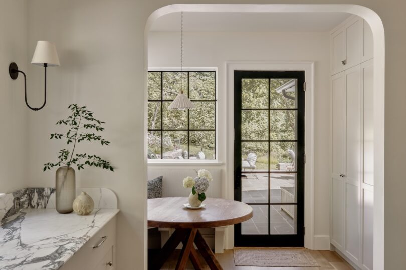 Bright kitchen nook with a round wooden table, vase of flowers, built-in seating, black-framed glass door, and large windows overlooking a green outdoor area.