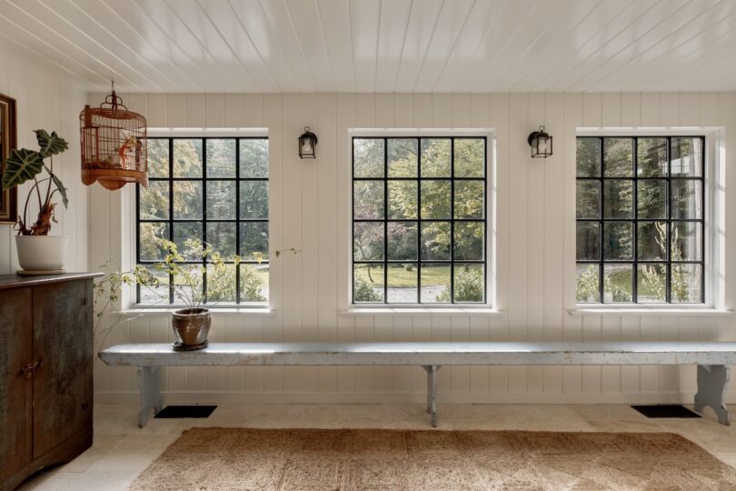 A bright room with three large windows, a long blue bench, a wooden cabinet, a hanging birdcage, potted plants, and outdoor scenery visible through the windows.