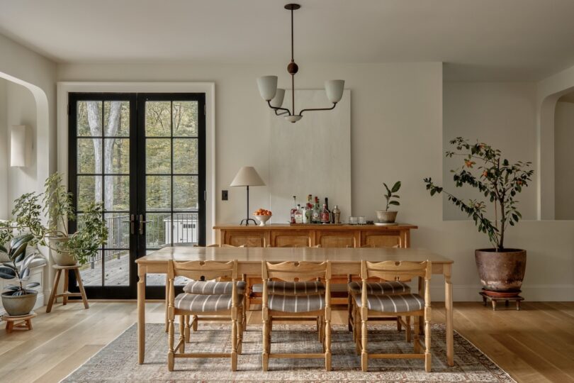 A dining room with a wooden table, six chairs, a sideboard with decor, potted plants, a rug, large windows, and a modern chandelier.