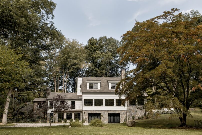 Two-story house with stone and wood exterior, large windows, and a steep roof, surrounded by trees and green lawn on a clear day.