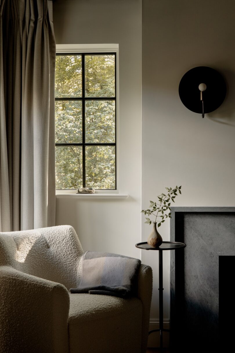 A cozy living room corner with a textured armchair, a small side table holding a vase with greenery, a window with trees outside, and a modern wall sconce above a dark fireplace.