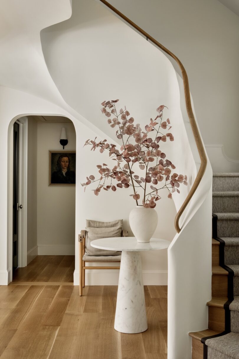 A modern hallway with light wood flooring, a marble pedestal table holding a white vase with pink foliage, a minimalist chair, and a curved staircase with a wooden handrail.