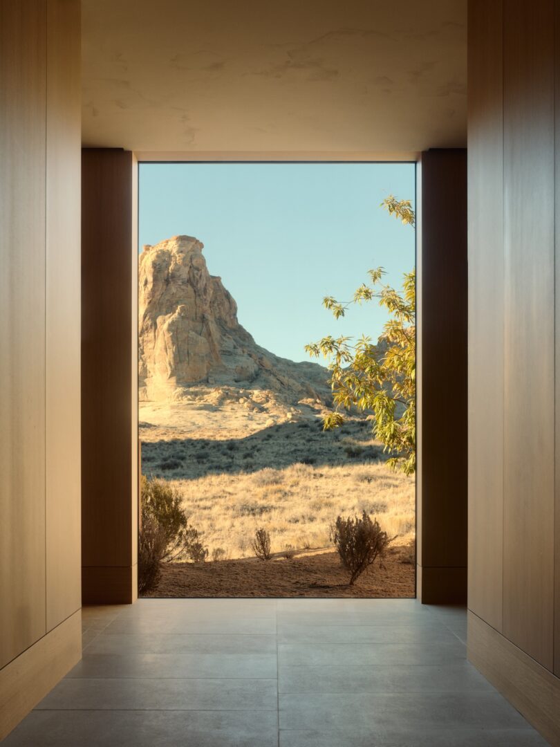 A large window in a modern, wood-paneled hallway frames a view of a rocky desert landscape with dry grass, shrubs, and a clear sky.