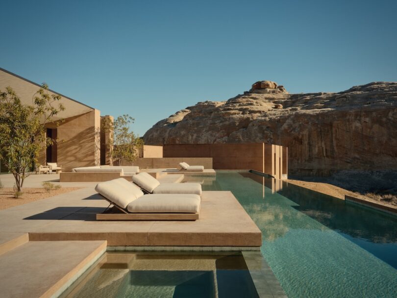 Modern outdoor pool with lounge chairs beside a minimalist building, set against a rocky desert landscape under a clear blue sky.