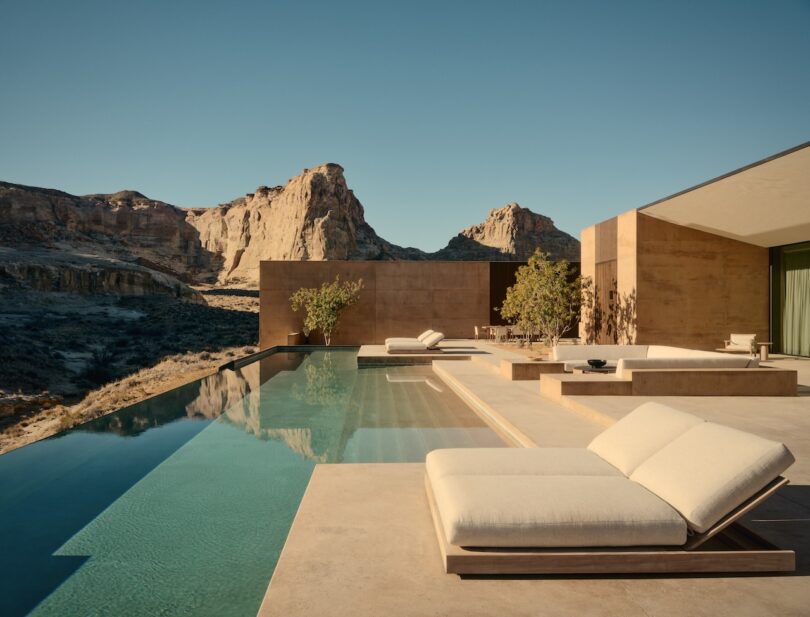 Modern villa with an infinity pool and lounge chairs overlooks a rocky desert landscape with large cliffs under a clear blue sky.