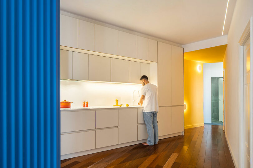 A man stands at a modern white kitchen counter with built-in cabinets, preparing food. The space features wood flooring, yellow accents, and a bright blue textured wall in the foreground.