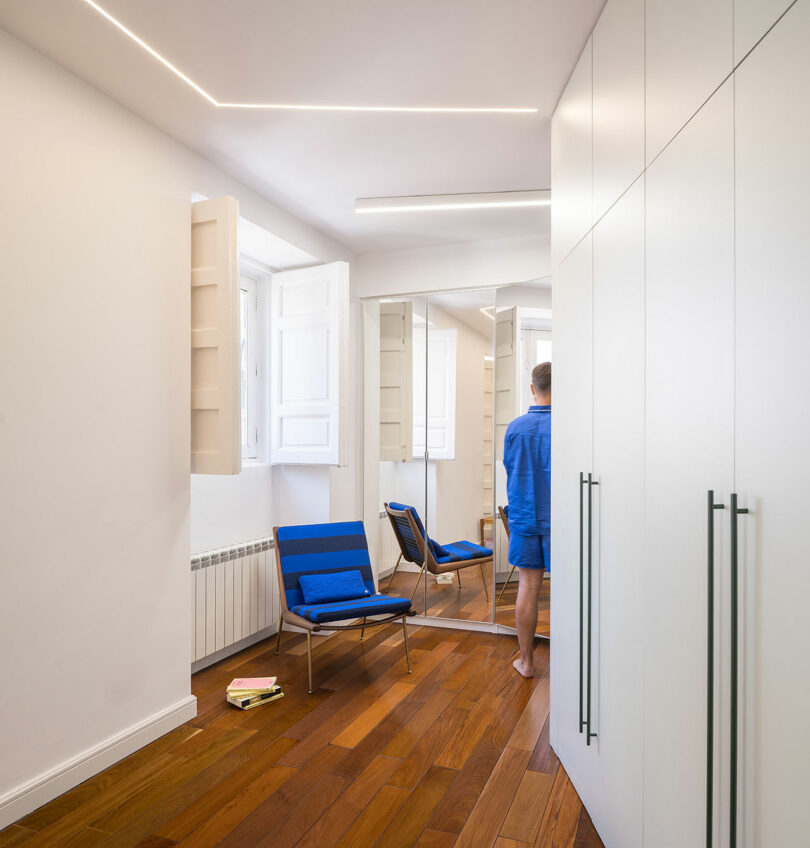 A person in blue stands by a mirror in a minimal, modern room with wood flooring, a blue chair, white cabinets, and an open window; books lie on the floor.