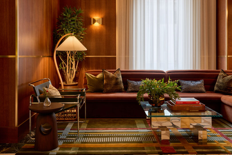 Modern living room by LEGEARD STUDIO with wood-paneled walls, a brown sectional sofa, glass coffee table, potted plants, books, and a patterned rug beneath a large window with sheer curtains.