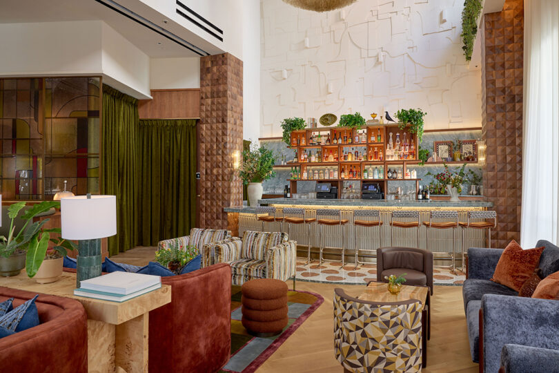 Modern hotel bar by LEGEARD STUDIO with patterned armchairs, a long bar counter with stools, shelves of bottles, and textured wall décor; plants and soft lighting create a warm, inviting atmosphere.