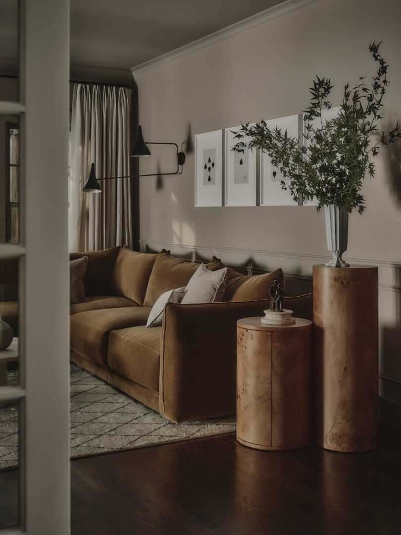 A modern living room with a brown velvet sofa, round wooden side tables, a vase with green branches, framed art on the wall, and floor-length curtains.