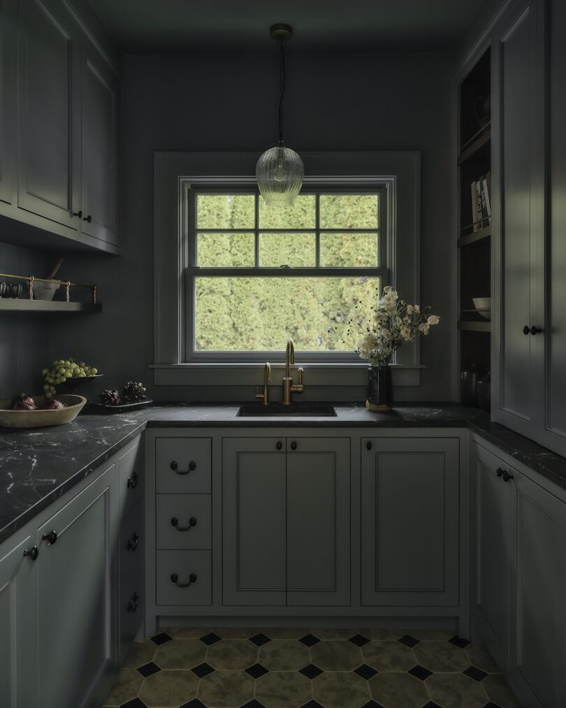 A small, dark-toned kitchen with black countertops, gold faucet, a window overlooking greenery, pendant light, and a vase of flowers by the sink.