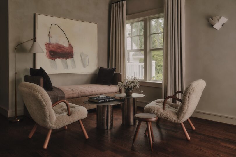 A cozy living room with two plush chairs, a wooden bench, a small round table, a stool, a large abstract painting, and a window with light curtains.
