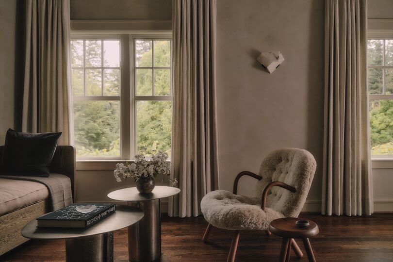 A cozy living room with large windows, beige curtains, a plush armchair, a small side table, a sofa, and a modern wall decoration; trees visible outside.