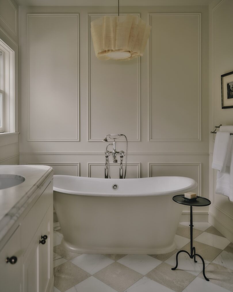 A freestanding bathtub sits in a neutral-toned bathroom with paneled walls, a window, a ceiling light, a towel rack, and a small black side table holding soap.