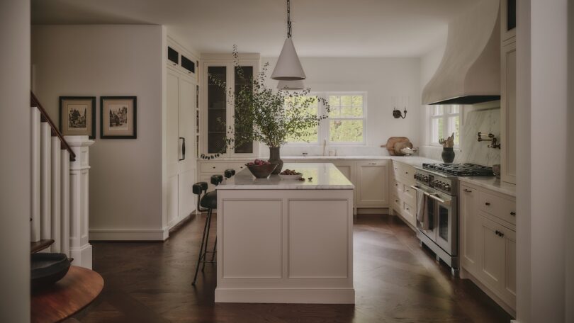 A modern kitchen with white cabinetry, marble island, bar stools, pendant light, wood floors, and large windows letting in natural light.