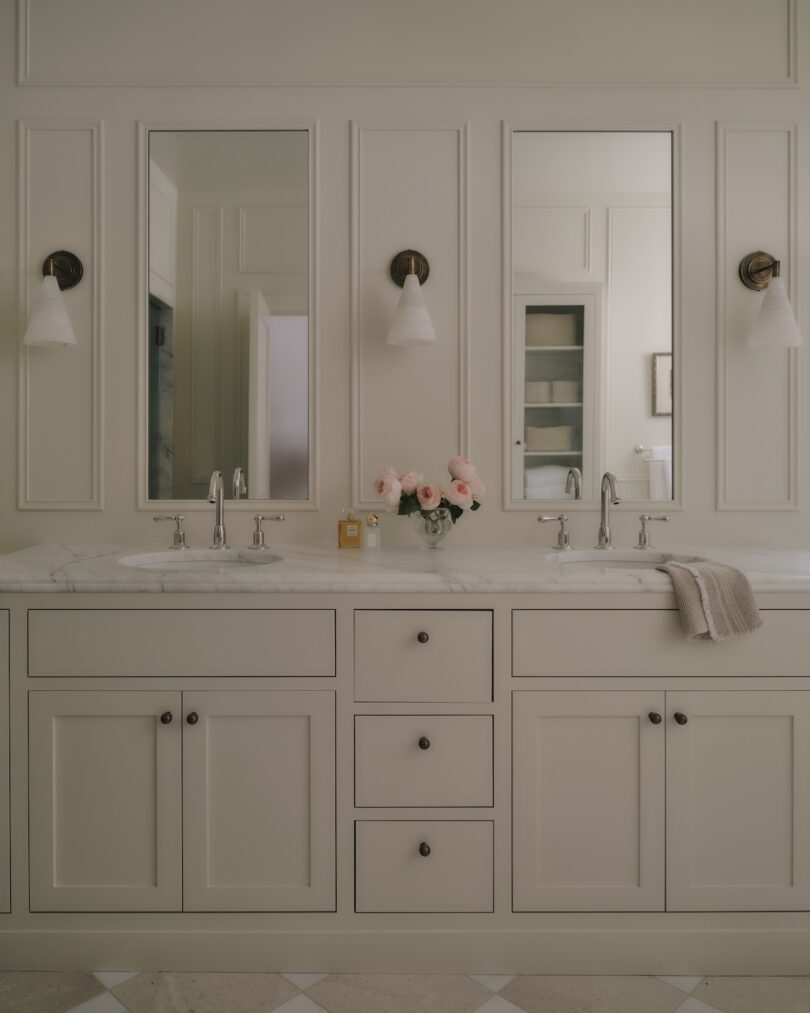 Double bathroom vanity with white cabinets, marble countertop, two mirrors, twin sconces, and a vase of pink flowers between two sinks. A hand towel is draped on the right.