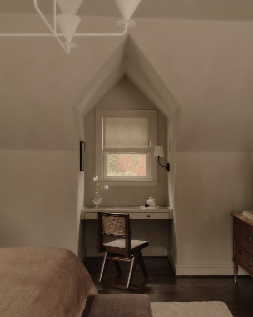 A small desk with a chair is placed in an alcove under a window with a beige shade; a single wall sconce, vase with flowers, and teapot sit on the desk.