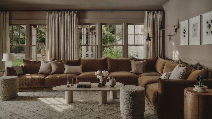 A spacious living room with a large brown sectional sofa, round coffee table, two textured stools, wall art, and floor-to-ceiling windows with beige curtains.