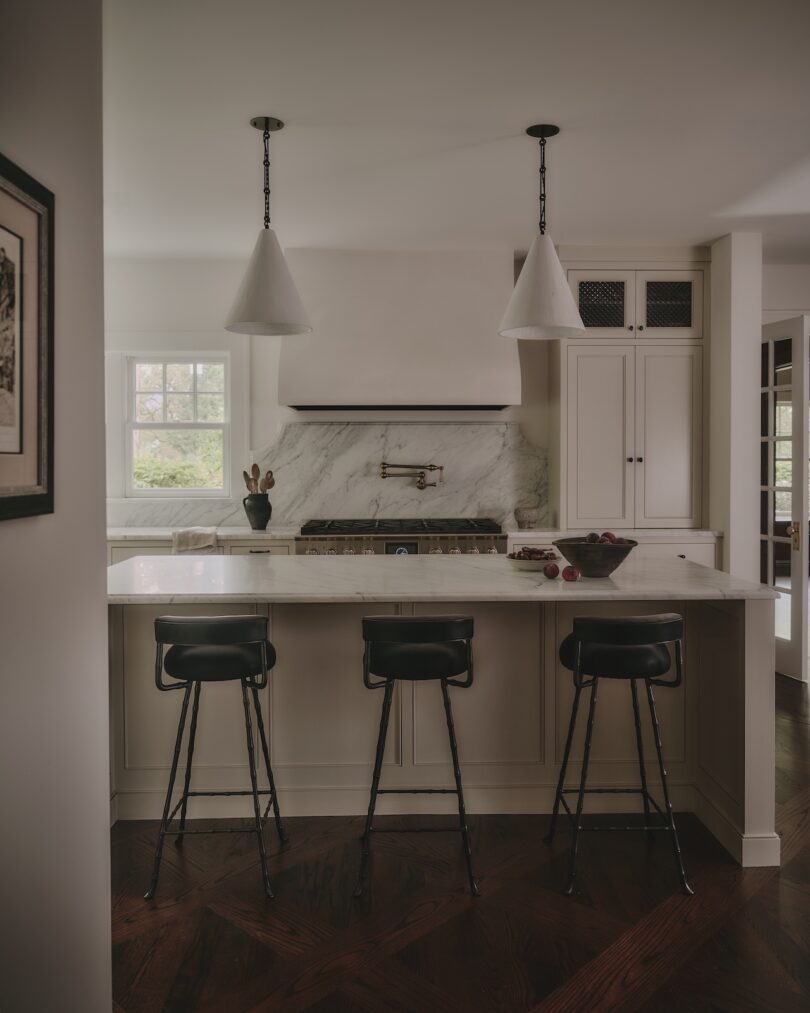 Modern kitchen with marble countertops, black barstools, pendant lights, and white cabinetry; a bowl and vase are on the island.