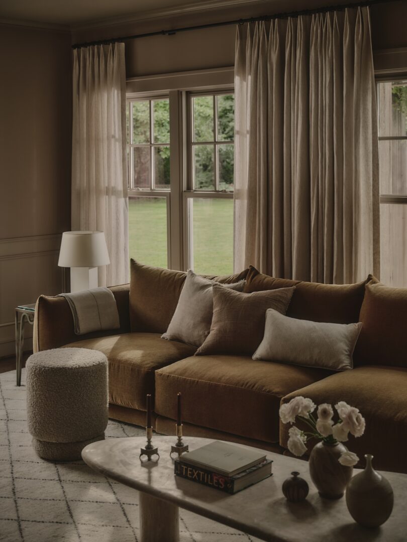 A living room with a brown velvet sofa, light cushions, a round textured ottoman, a marble coffee table with candles, flowers, and books, and large windows with beige curtains.