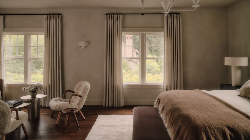 A cozy bedroom with two windows, neutral walls, a bed with brown bedding, beige curtains, two plush chairs, a small table with flowers, a lamp, and a white rug on wood flooring.