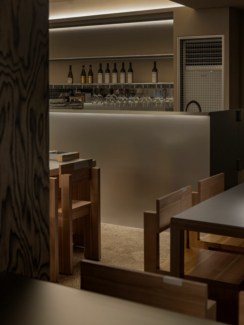 Minimalist bar interior with wooden chairs and tables, soft lighting, glassware on the counter, and bottles arranged on a shelf behind the bar.