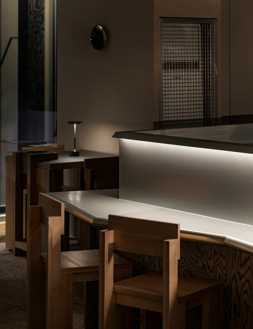 A modern, dimly lit interior with wooden chairs and tables, a sleek illuminated counter, and minimalistic decor.