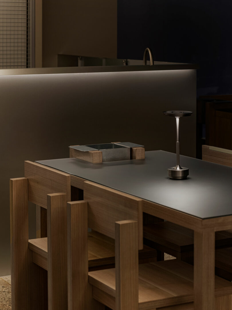 A modern wooden dining table with matching chairs, a square tray, and a small lamp on a dark tabletop in a minimalist interior setting.
