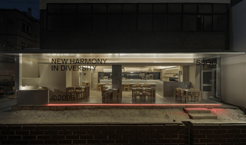 A modern, glass-fronted restaurant with minimalist wooden furniture and the text "NEW HARMONY IN DIVERSITY SSOC ©2025" displayed on the window at night.