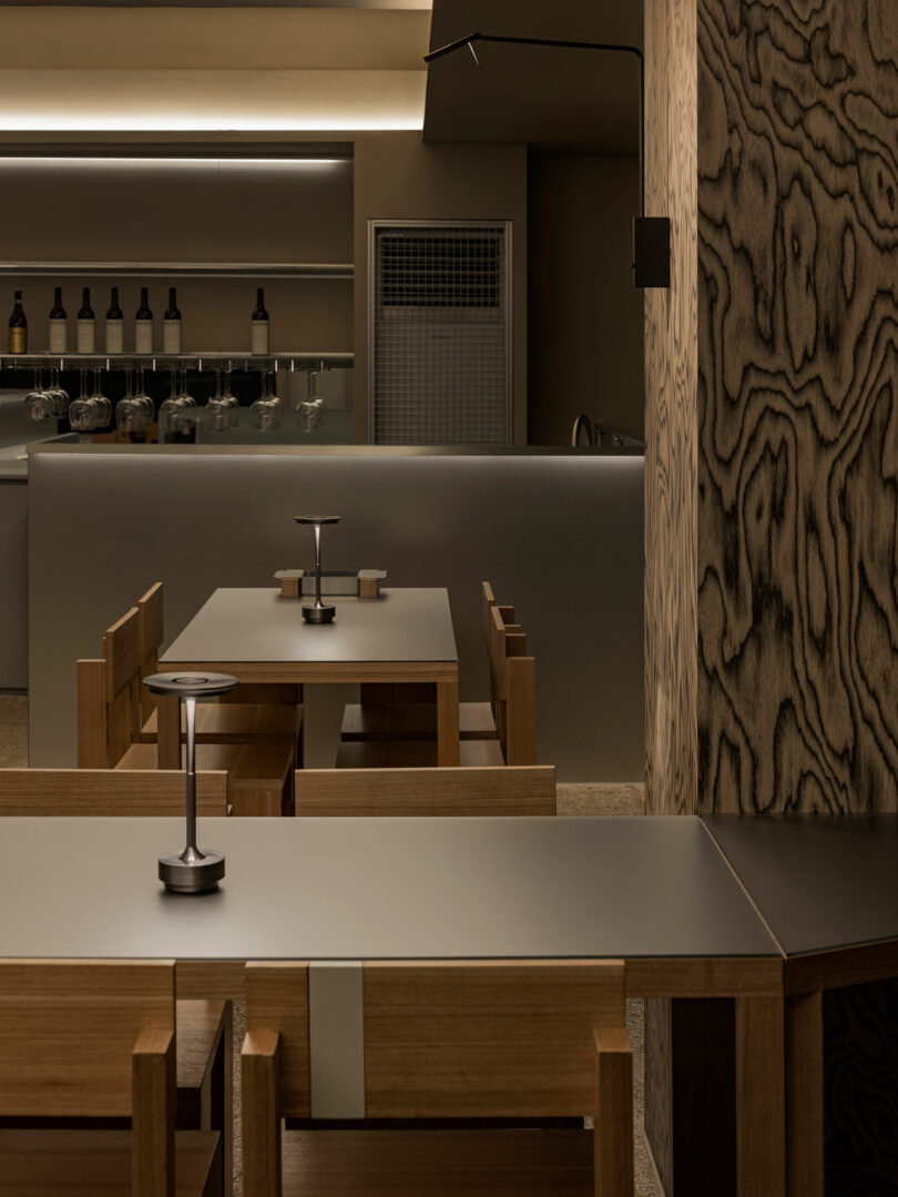 Modern minimalist dining area with wooden tables and chairs, soft lighting, and a sleek bar counter in the background.
