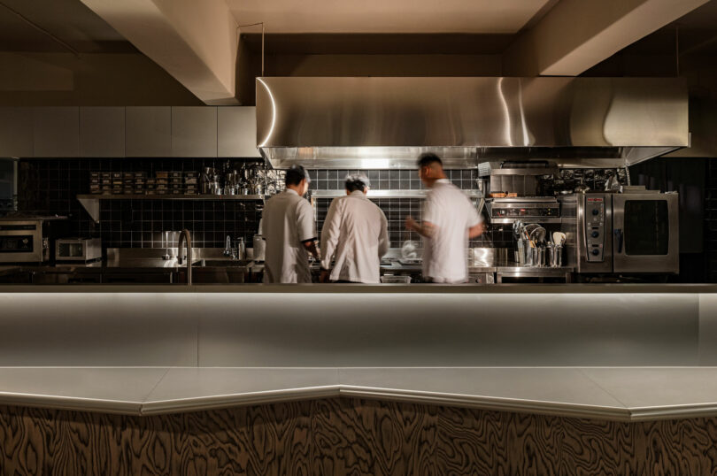 Three chefs in white uniforms work in a modern, stainless steel restaurant kitchen with various appliances and utensils visible.